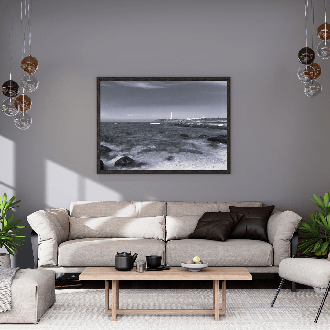 A framed black and white coastal landscape photograph hangs on a gray wall above a light beige sofa in a modern living room setting.