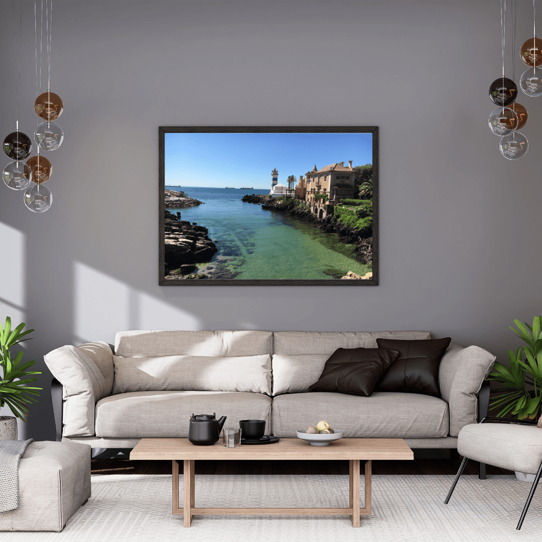 A framed landscape art print of a coastal town with turquoise water and a lighthouse, displayed on a gray wall above a modern beige sofa in a styled living room.