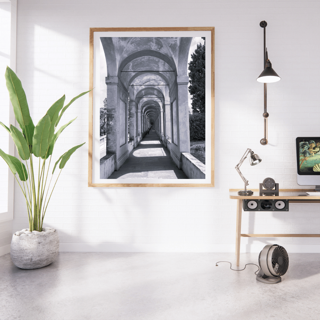 A framed black and white architectural art print depicting a stone archway corridor, displayed on a white wall in a modern, sunlit home office setting with a desk, lamp, and potted plant.