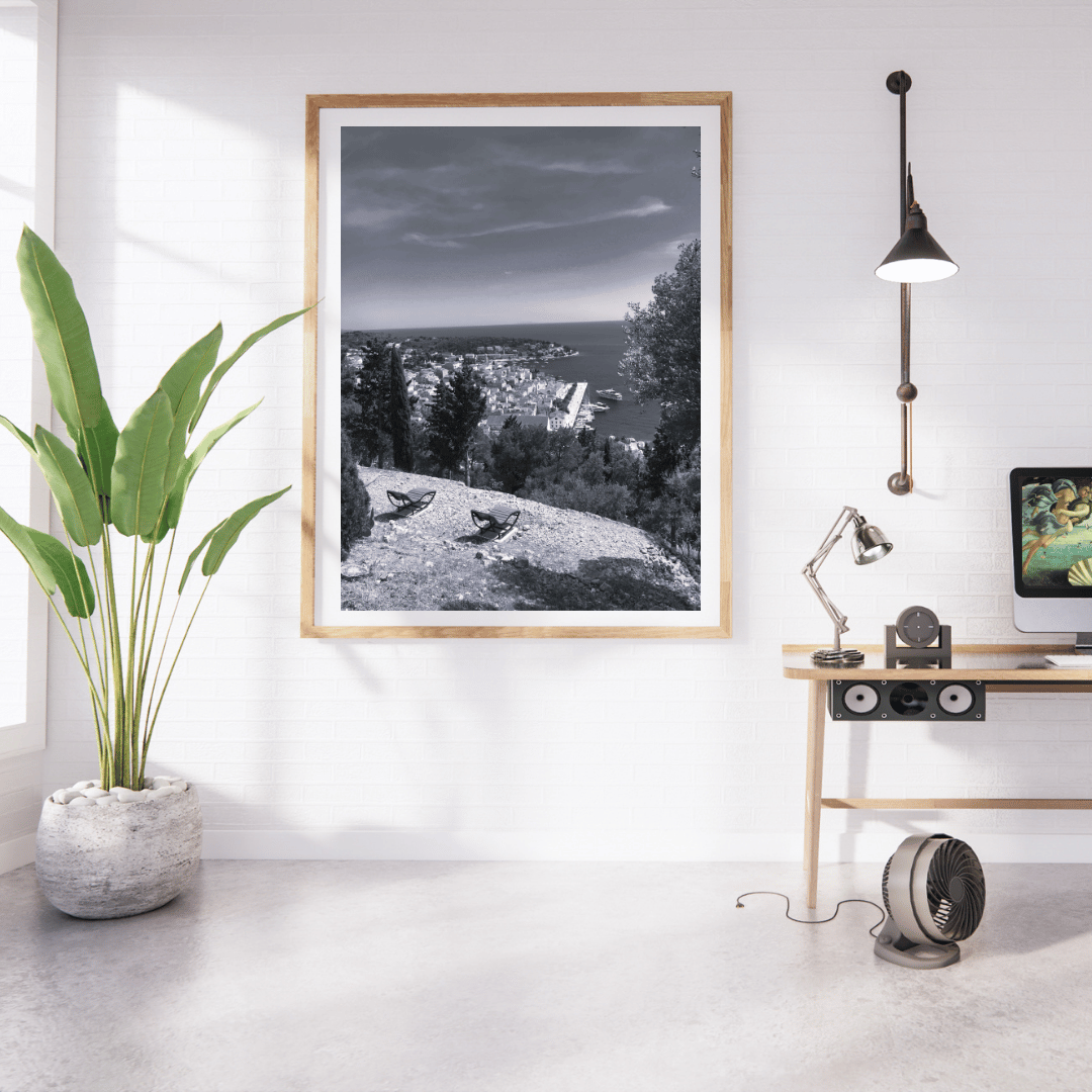 A framed black and white landscape photograph of a coastal town hanging on a white wall in a modern, sunlit room with a plant, desk, lamp, and fan.