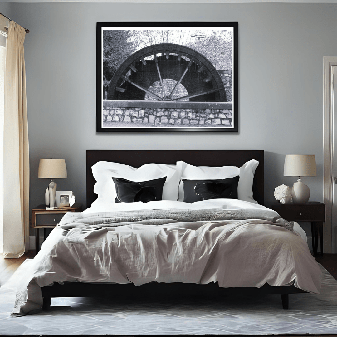 A black and white framed wall art print depicting a rustic water wheel in a regional small village in Provence, the south of France, displayed above a neatly made bed in a modern, neutral-toned bedroom.