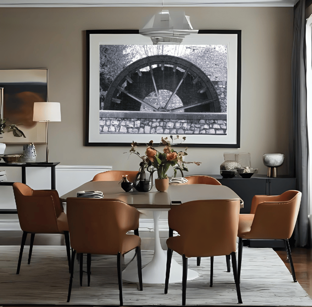 A black and white framed wall art print depicting a rustic water wheel in a regional small village in Provence, the south of France, hangs on a beige wall in a modern dining room setting with a wooden dining table and six brown upholstered chairs.