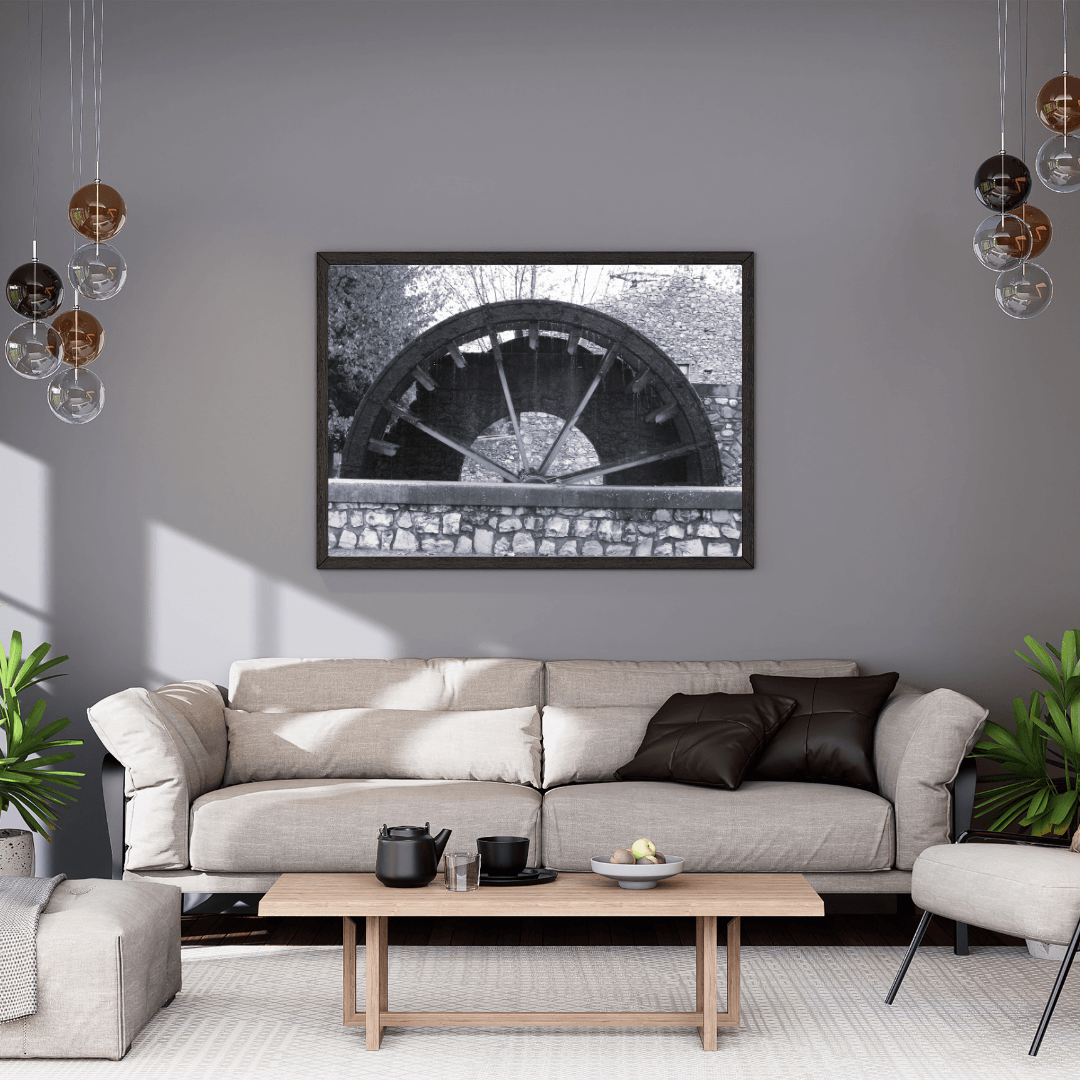 A black and white framed wall art print depicting a rustic water wheel in a regional small village in Provence, the south of France, hanging on a gray wall above a modern beige sofa in a styled living room.