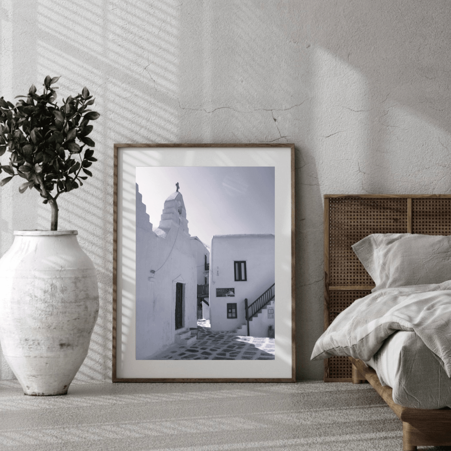 A framed black and white architectural art print leaning against a textured white wall in a modern bedroom setting, next to a bed and a large white vase with a potted plant.